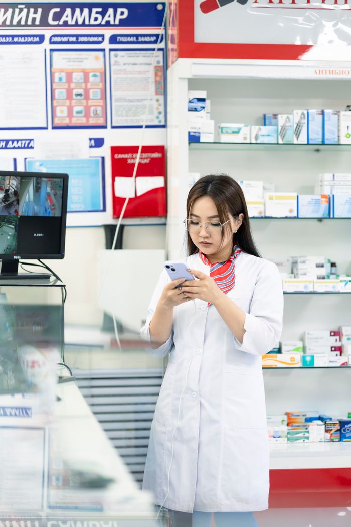 A female pharmacist in a lab coat using a smartphone in a pharmacy, surrounded by medicine shelves.
